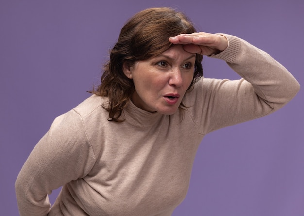 Free Photo middle aged woman in beige turtleneck looking far away with hand over head standing over purple wall