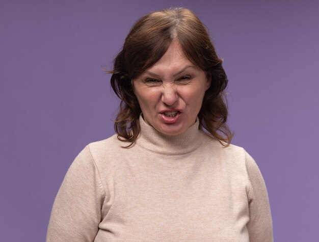 Middle aged woman in beige turtleneck  with angry face standing over purple wall