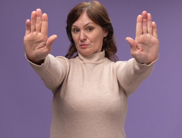 Middle aged woman in beige turtleneck  with serious face making stop gesture with hands standing over purple wall