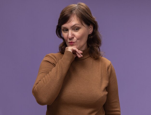 Middle aged woman in brown turtleneck looking aside with hand on chin thinking positive standing over purple wall