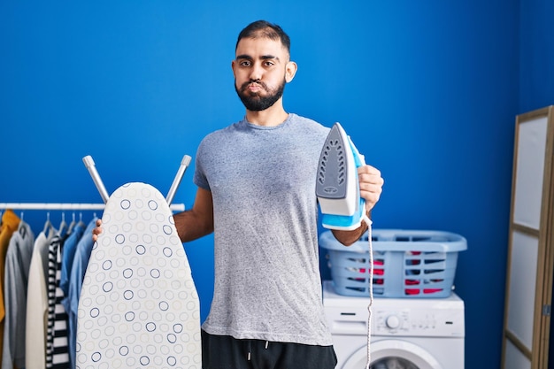 Free photo middle east man with beard holding electric iron puffing cheeks with funny face mouth inflated with air catching air