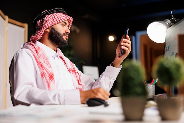 Middle Eastern businessman dressed traditionally sits and holds his smartphone at a desk in his office Arab guy grasping a mobile device and using the desktop computer nearby