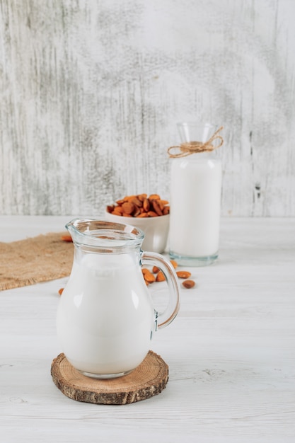 Free photo milk carafe with bowl of almonds and bottle of milk high angle view on a white wooden and piece of sack background