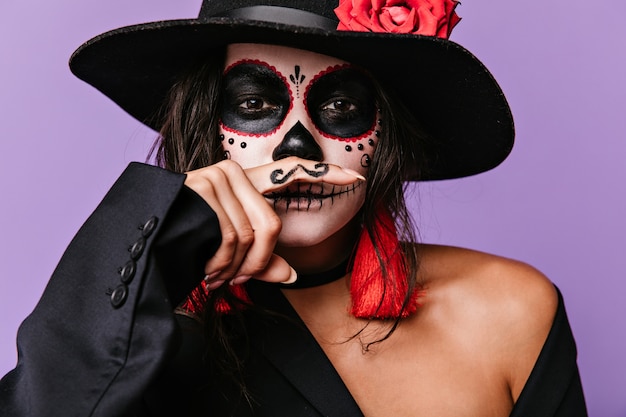 Mischievous brown-eyed girl with face art is having fun by putting painted mustaches to her mouth. Photo of stylish woman in black outfit with red details.