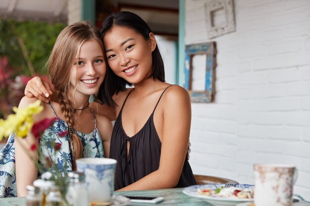 Mixed race beautiful young women of different nationalities embrace as demonstate interracial true friendship or relationships, drink coffee in bar