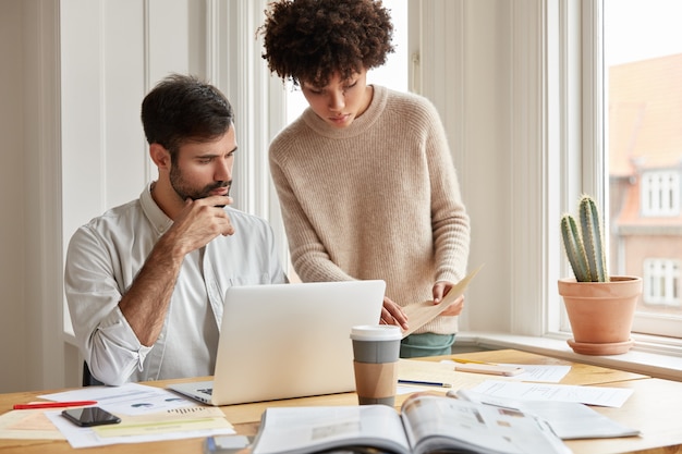 Mixed race family couple examine paperwork, ask advice each other, pose against cozy domestic interior near window, use laptop computer, drink takeaway coffee.