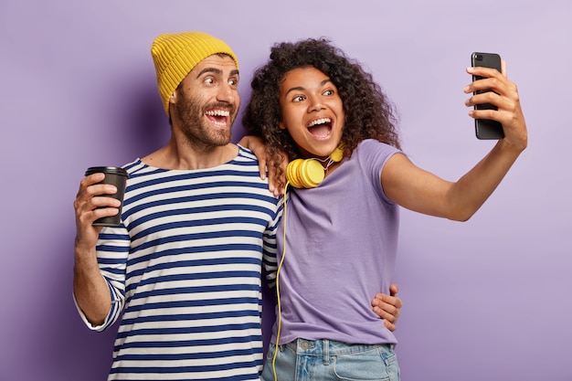 Free photo mixed race lovely couple embrace and stand closely, pose for making selfie portrait, have glad expressions, drink coffee