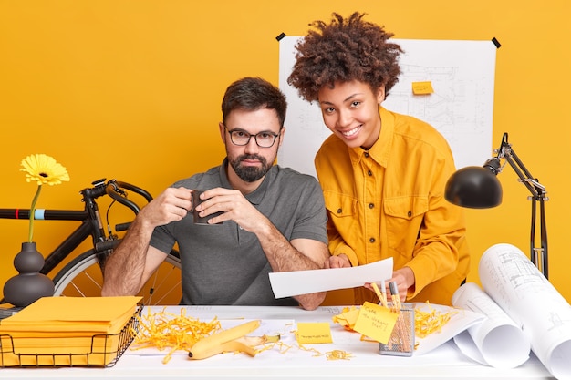 Mixed race woman and man collaborate together discuss future project and try to find best variant for sketches pose at messy desktop 