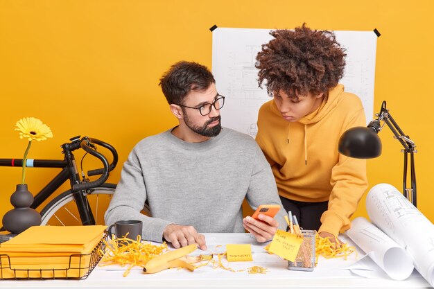 Mixed race woman and man coworkers try to find solution of work check information in internet via mobile phone study papers pose in coworking space enjoy cooperation 