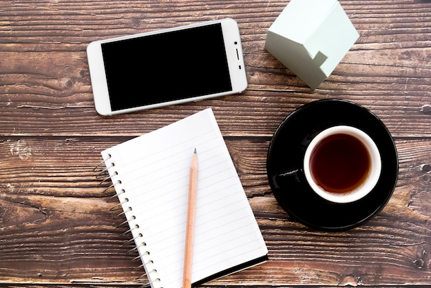 Mobile phone; blank spiral notebook; pencil; coffee cup and house model on wooden desk