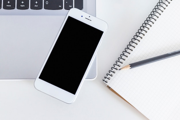 Mobile phone over the laptop with pencil and notebook over the white table