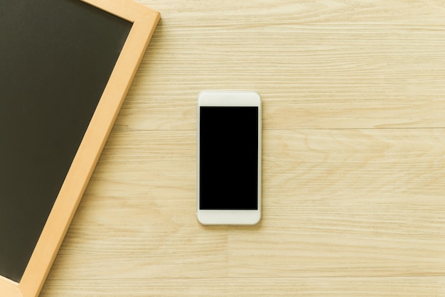 Mobile phone with blank screen and blank wooden blackboard frame on wooden table background. Top view with copy space. Can be used mock up image. Vintage effect style pictures.