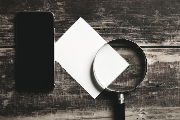 Mobile smartphone, magnifier and sheet of white paper isolated on black farm wooden table