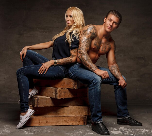 Modern fashionable couple. Shirtless tattoed man and sensual blond woman sitting on wooden blocks.