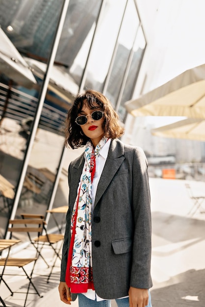 Modern fashionable woman with shirt hairstyle in sunglasses with red lips in grey jacket and bright silk scarf is posing outside on sunny street