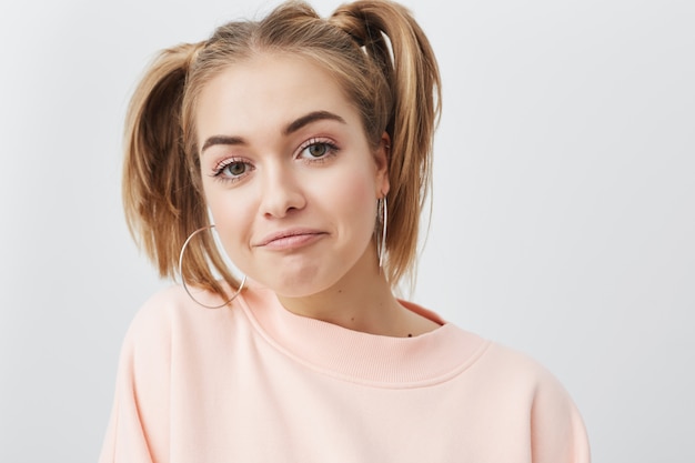 Modern generation, youth and happiness concept. Picture of funy stylish student girl with two ponytails, spending her leisure time indoors, in pink sweatshirt and with big round earings.