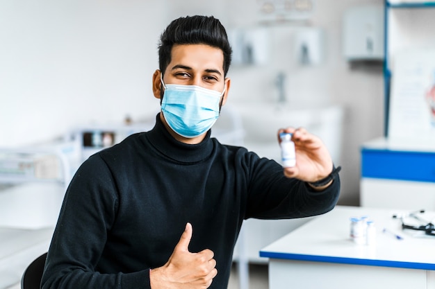 A modern Indian man holds a vaccine in his hands and smiles, wearing a protective mask.