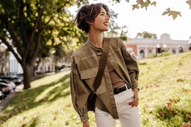 Modern lady in denim jacket with handbag looking away outside. Wavy-haired woman with red lips in white pants smiling outdoors.