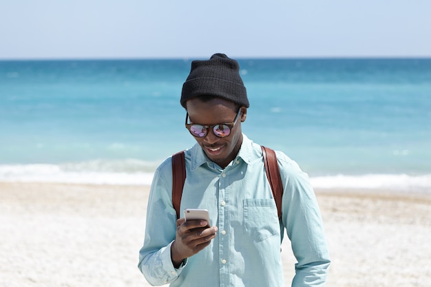 Modern lifestyle, technology and communication concept. Attractive young black European hipster in shirt, hat and sunglasses enjoying high-speed 3g or 4g internet while spending weekend by the sea