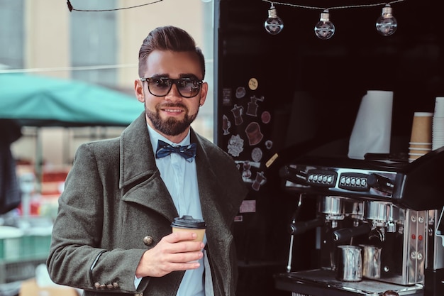 Free photo modern smiling man in sunglasses is drinking coffee while standing outside at coffeeshop.