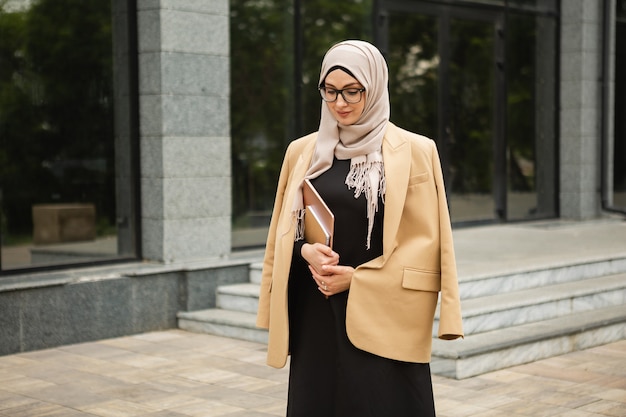 Free photo modern stylish muslim woman in hijab, business style jacket and black abaya walking in city street with laptop