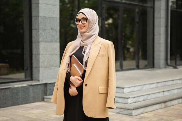 Modern stylish muslim woman in hijab, business style jacket and black abaya walking in city street with laptop