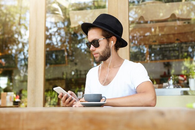 Modern technology and communication concept. Handsome student in white earphones relaxing at outdoor coffee shop making video call