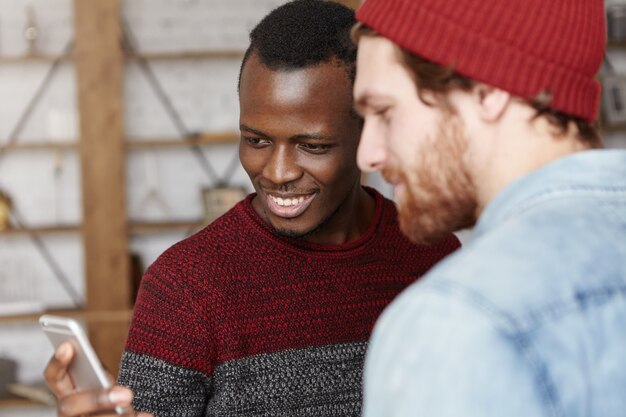 Modern technology, people, leisure and online communication. Happy young dark-skinned male holding cell phone and showing something to his stylish Caucasian friend, both looking at screen and smiling