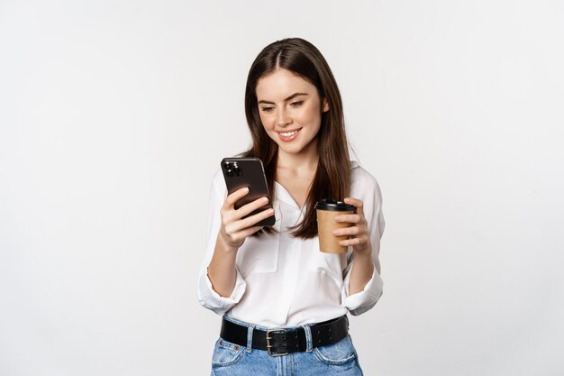 Modern woman looking at mobile phone and drinking from takeaway cup coffee standing over white backg...