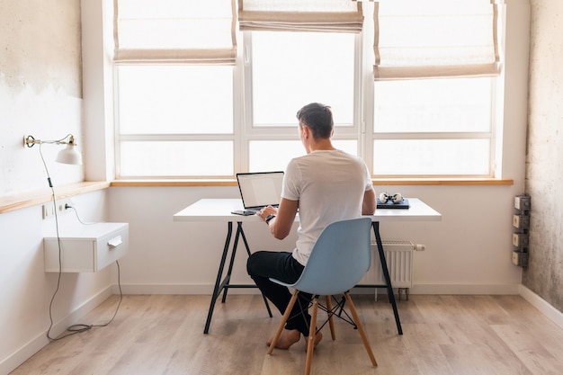 Free photo modern young handsome man in casual outfit sitting at table working on laptop, freelancer at home