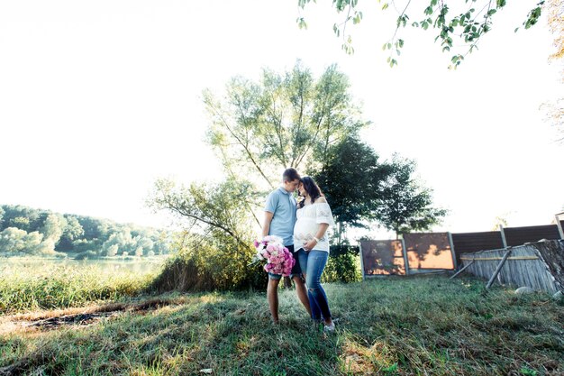 Modish expecting couple stands in warm embraces under green tree