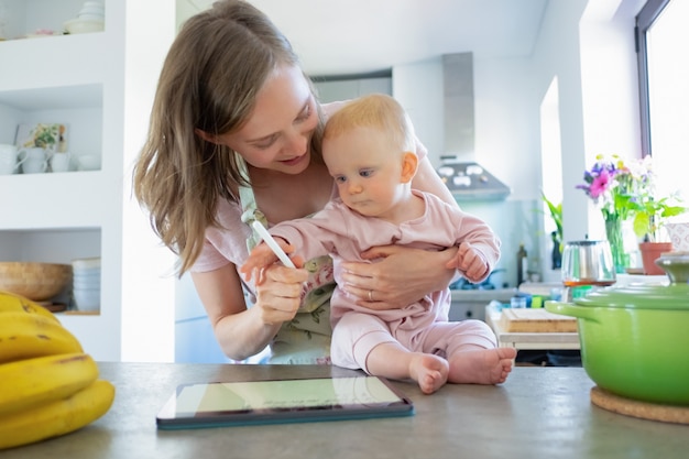 Free photo mom and baby daughter cooking together at home, watching recipes on tablet. child care or cooking at home concept