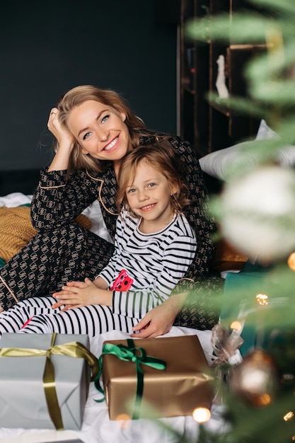 Free photo mom and daughter are sitting in pajamas on the bed by the christmas tree at christmas new year famil