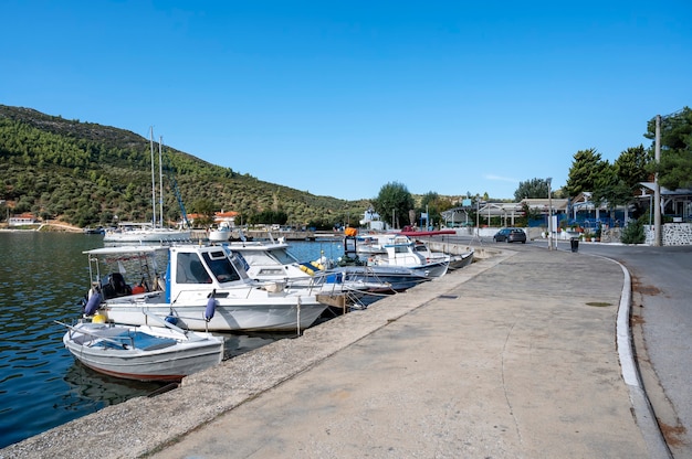 Moored boats on the water near the embankment street with buildings and restaurants, a lot of greenery, green hills, Greece