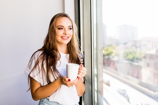 Morning coffee is my daily routine. Young woman drink cup of morning coffee near windows at home looking at street. Morning mood.