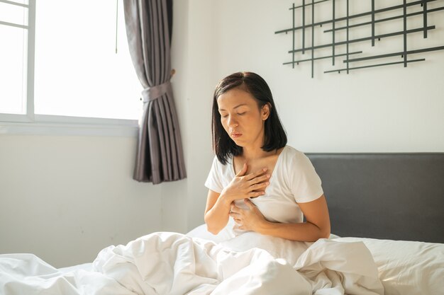 Morning sickness. Young pregnant woman sitting on bed, covering her mouth feeling nauseous during pregnancy, Woman in white pajamas suffering from Acid reflux while wake up on her bed in the morning.