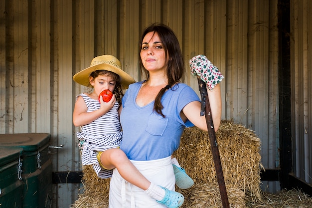 Free Photo mother carrying her daughter eating red apple standing in front of hay stack