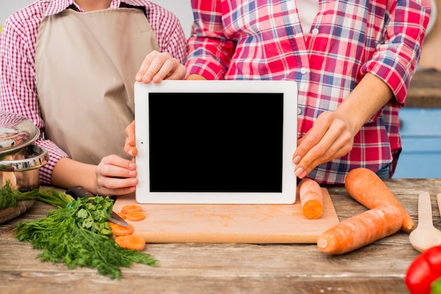 Free Photo mother and daughter showing blank screen digital tablet on chopping board with vegetables