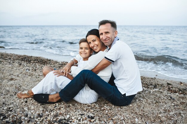 Mother, father and son is sitting on the beach near the sea, hugging and looking straight