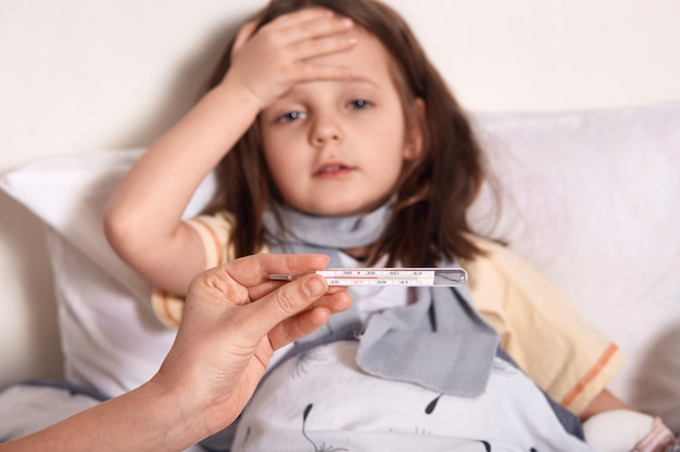 Mother holding thermometer in hands, sick little girl lying in bed and keeping her palm on forehea, suffering from high temperature