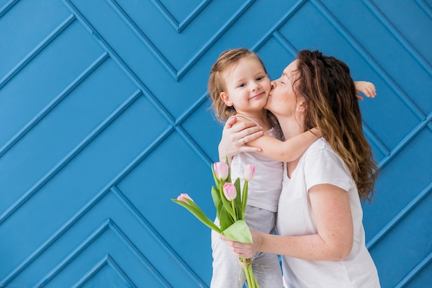 Mother kissing to her pretty little daughter holding tulip flowers over blue background