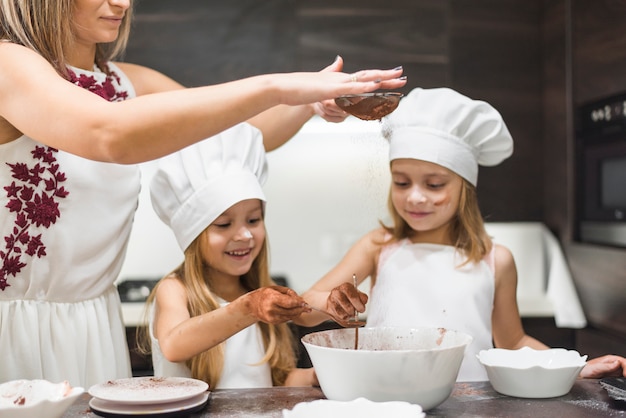 Free photo mother sifting cocoa powder with strainer in bowl while happy girls preparing food