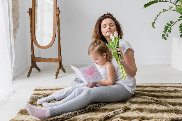 Free photo mother smelling fresh tulip flowers while daughter reading greeting card sitting on soft carpet