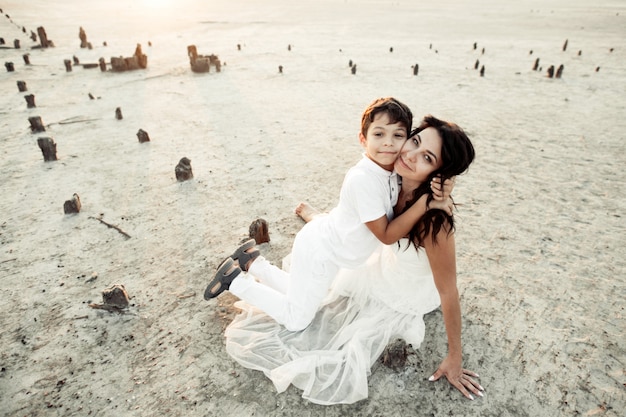 Free Photo mother and son is sitting on the sand dressed in white clothes, smiling and hugging