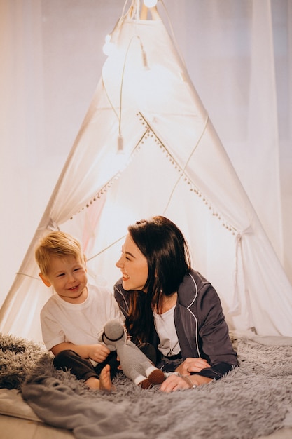 Mother with son sitting in cozy tent with lights at home on Christmas