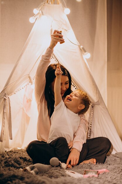 Mother with son sitting in cozy tent with lights at home on Christmas