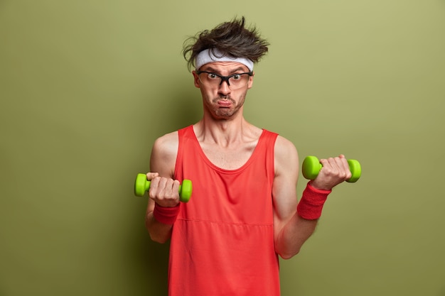 Motivated determined sportsman leads sporty lifestyle, lifts heavy dumbbells for muscle training, does morning fitness at home, wants to have biceps, wears red shirt and wristband, looks sadly