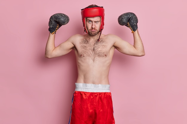 Motivated sportsman enjoys boxing wears protective hat gloves raises arms shows muscles has skinny body stands with bare torso looks seriously.