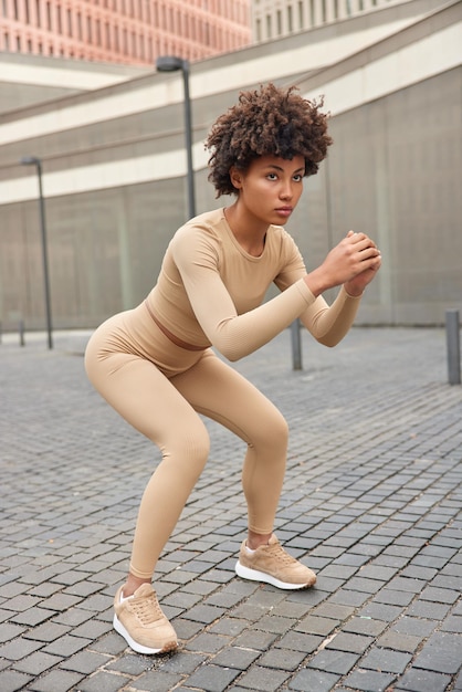 Free photo motivated sporty curly haired serious woman does deep squats in urban setting exercises outdoors for staying healthy and fit dressed in beige activewear sneakers focused forward workout concept