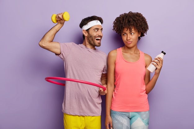 Free photo motivated young man exercises with hula hoop, raises dumbbell, has glad expression, wears white headband and t shirt and displeased woman stands with bottle of water, has fitness training